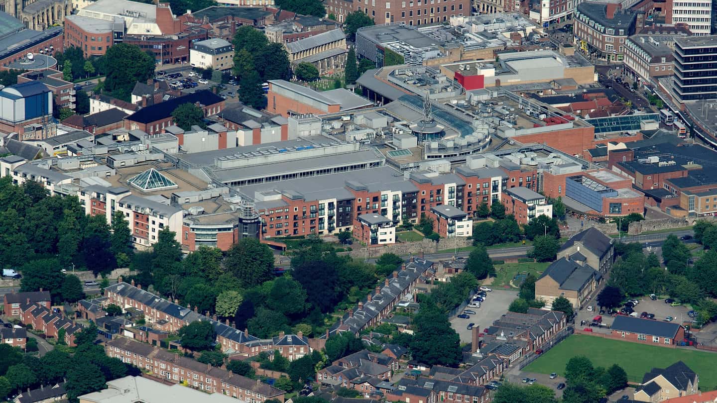 Aerial photograph of a city-centre retail block: a wide low-slope roof carrying rows of rooftop mechanical units and rooflights, ringed by brick apartment blocks and terraced streets.