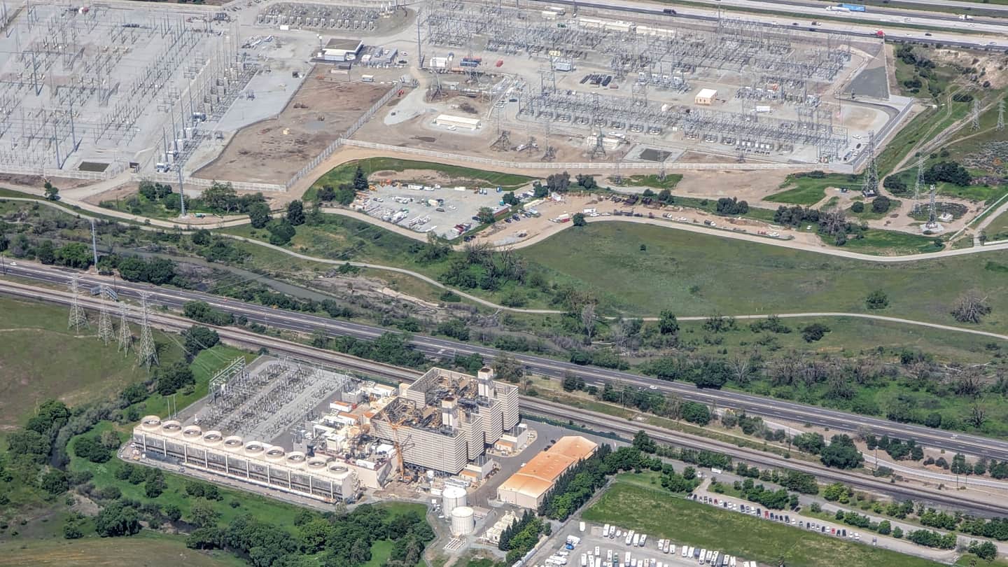 Aerial photograph of a high-voltage substation switchyard and its transmission towers, separated by a rail corridor and a freeway from a generating plant and a truck yard.