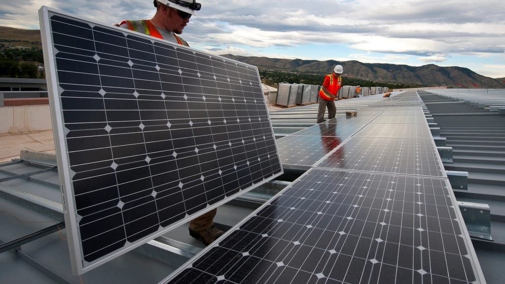 Workers part-way through fitting a photovoltaic array on a low-slope commercial metal roof: one panel stands on edge in the foreground and completed rows run away toward hills on the horizon.