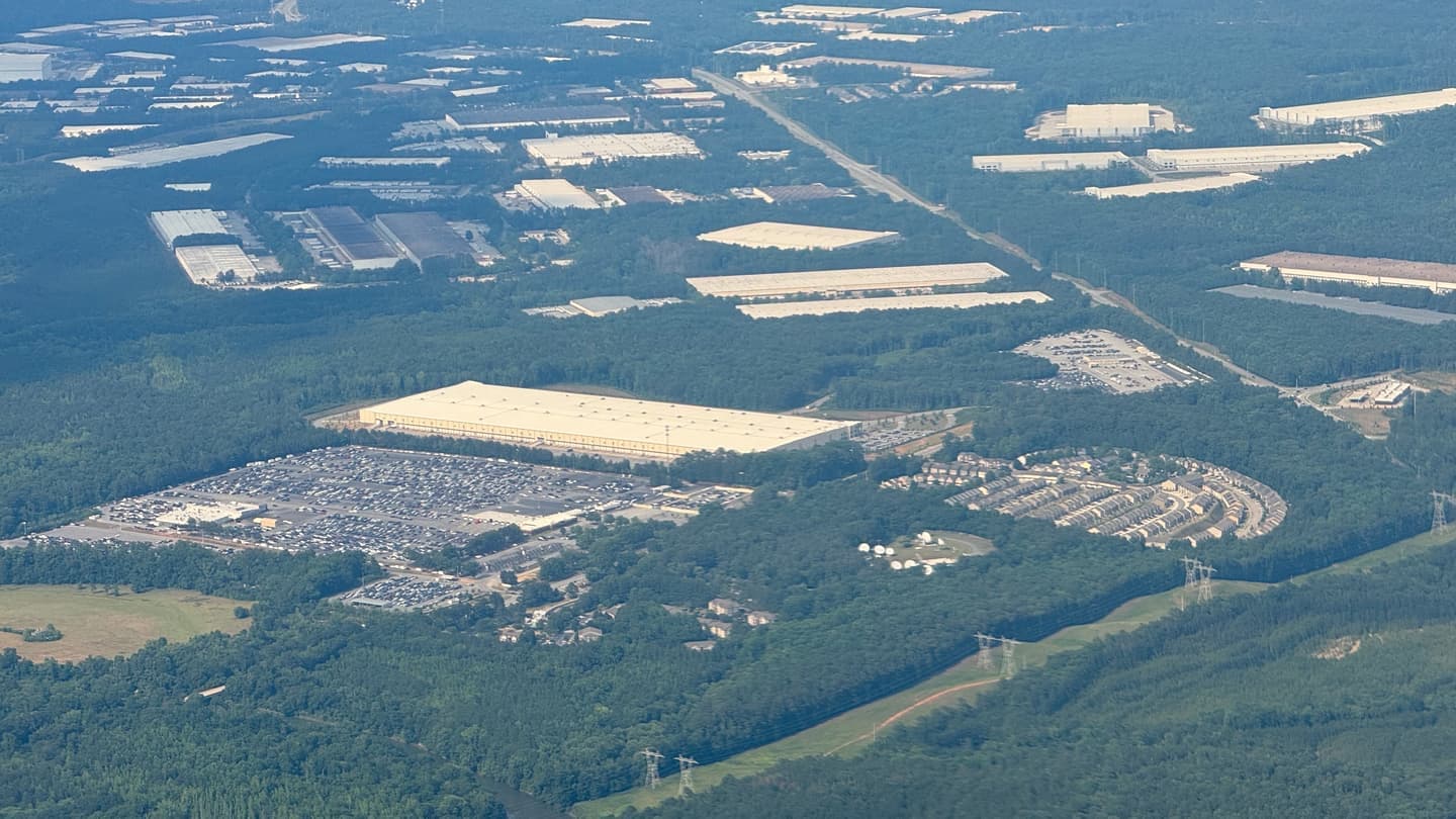 Aerial photograph of a warehouse district: one very large pale distribution-centre roof beside a full employee parking lot, with dozens of smaller flat industrial roofs and belts of woodland behind it.