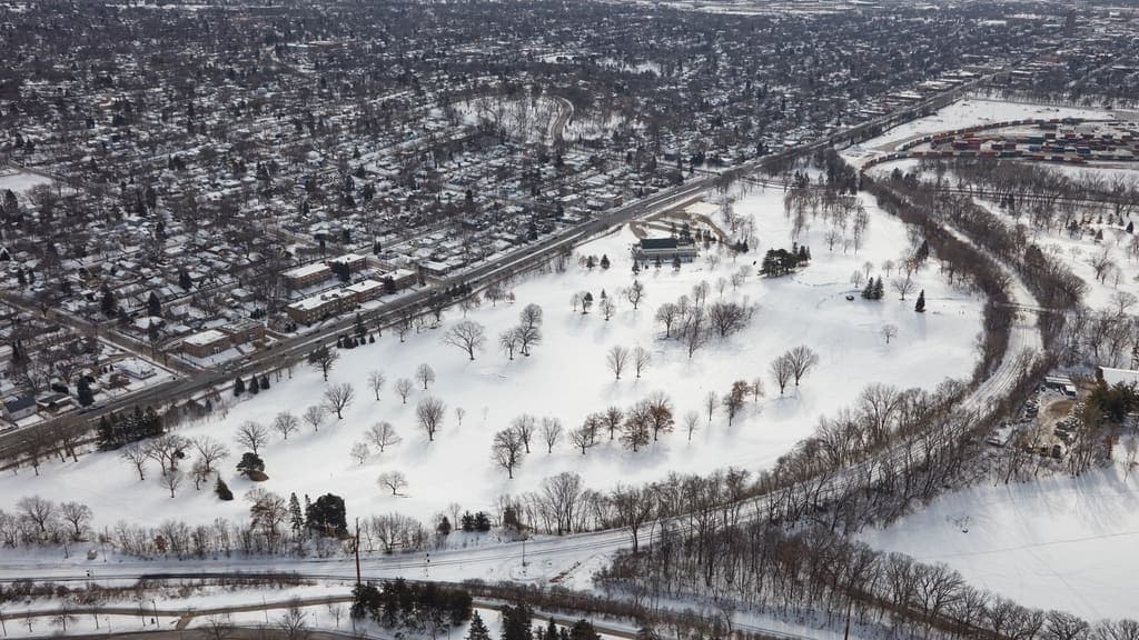 Aerial photograph of a snow-covered suburb in winter: a grid of residential streets and small commercial buildings on one side of an arterial road, and an open tree-lined park on the other.