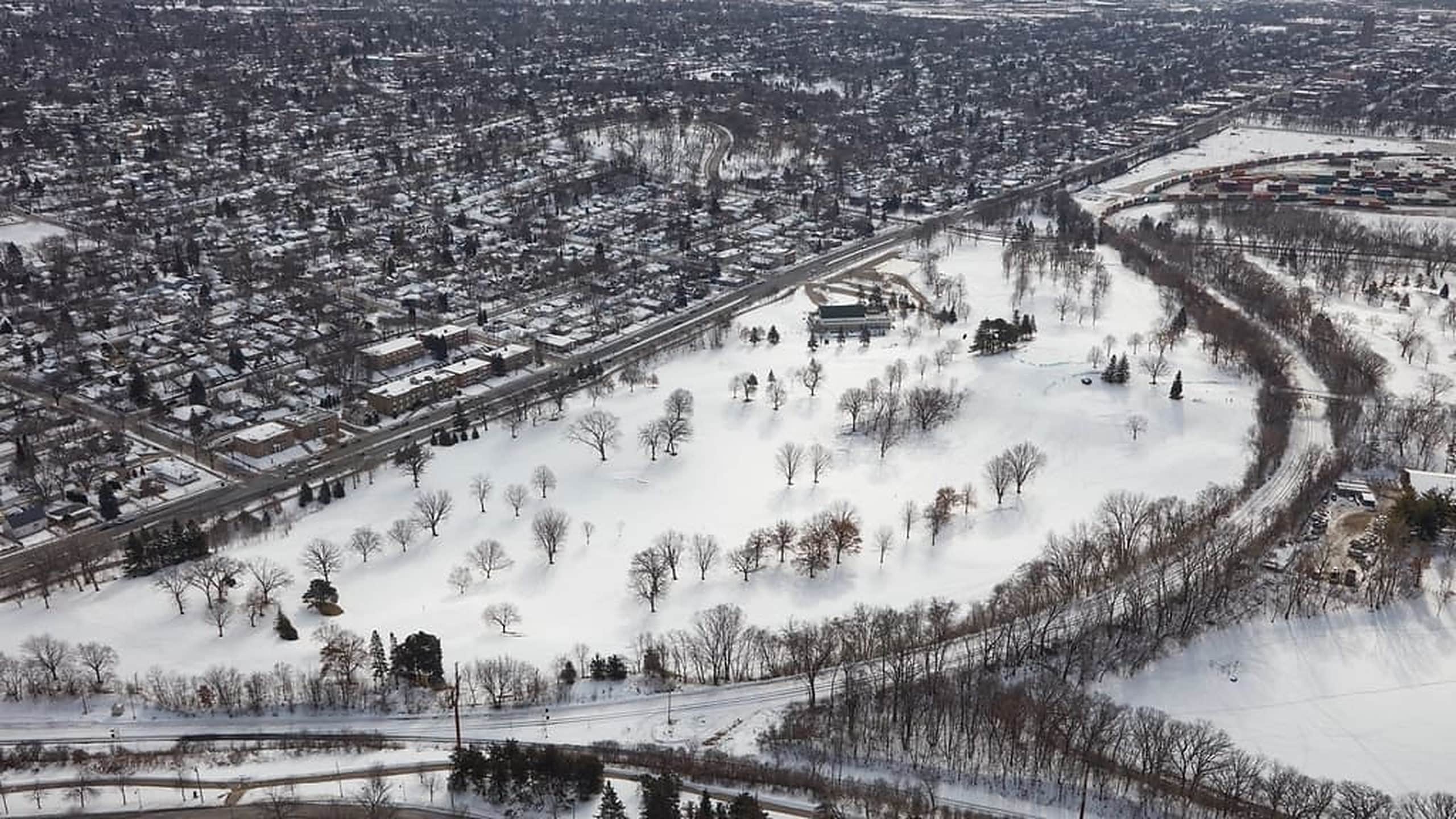 Aerial photograph of a snow-covered suburb in winter: a grid of residential streets and small commercial buildings on one side of an arterial road, and an open tree-lined park on the other.